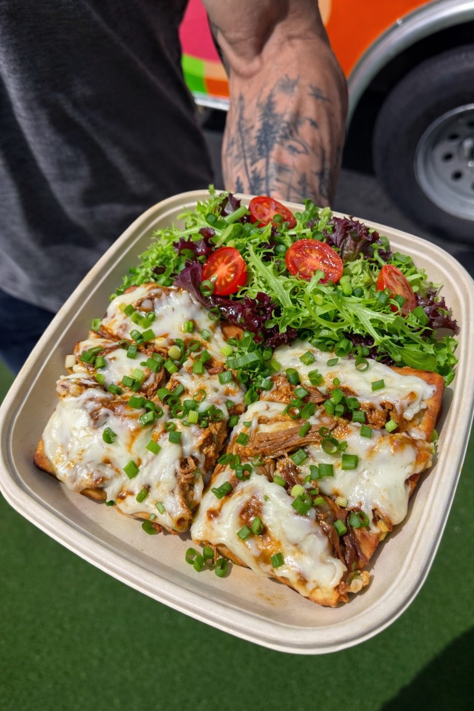 A person holds a takeout container with cheesy flatbread and a side salad with cherry tomatoes.